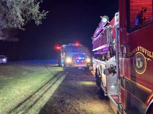 Two fire trucks with flashing lights are parked on grass at night. The side of one red truck reads “Sergentsville” while another emergency vehicle and a car are visible in the background.