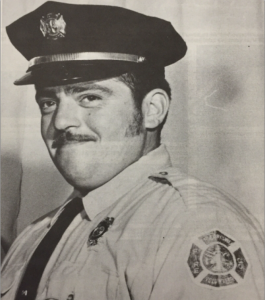 A man in a firefighter’s uniform and cap smiles slightly, looking into the camera. The uniform has badges and a patch with a fire department emblem on the sleeve. The image is black and white.
