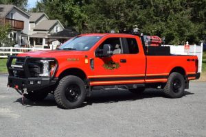 A bright red fire department brush truck is parked on a gravel road in a residential neighborhood, equipped with off-road tires and firefighting equipment in the truck bed.