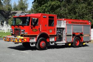 A bright red fire truck labeled "STEECHATTVILLE" and "ENGINE 47" is parked on a gravel surface with trees and a house in the background. The truck has shiny metal panels and emergency equipment attached.
