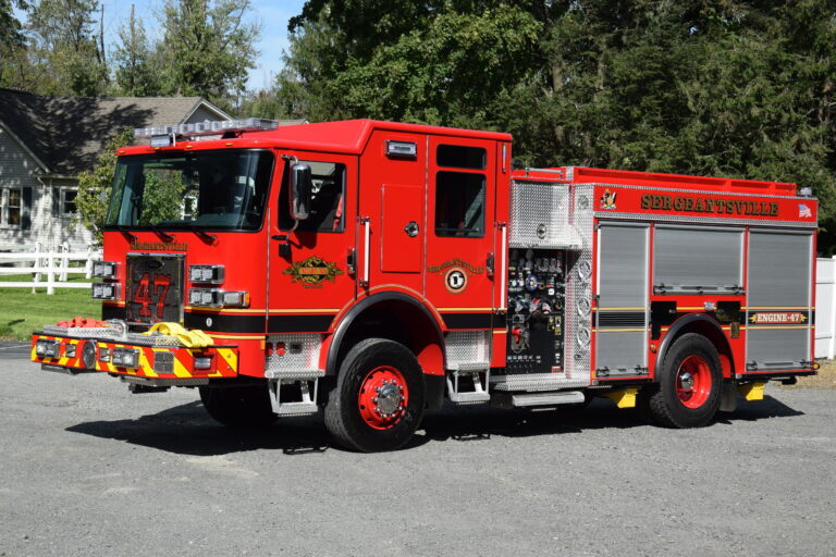 A bright red fire truck labeled "STEECHATTVILLE" and "ENGINE 47" is parked on a gravel surface with trees and a house in the background. The truck has shiny metal panels and emergency equipment attached.