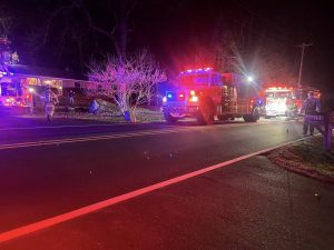 Two fire trucks with flashing lights are parked on a dark road at night in front of a house. Firefighters are visible near the vehicles, and the scene is illuminated by red and blue emergency lights.