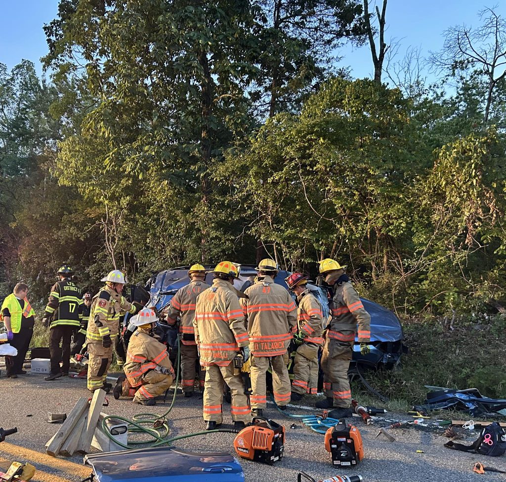 A group of firefighters in full gear work to rescue someone from a damaged SUV after a crash on a road beside trees. Tools and equipment are scattered on the ground, and other emergency personnel are nearby.