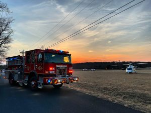A red fire truck is parked on the roadside at sunset, with emergency lights on. In the background, a medical helicopter and several vehicles are on a grassy field under a colorful sky.