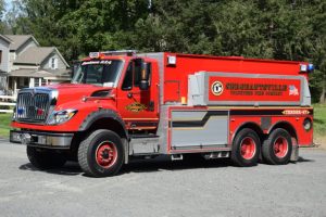 A bright red fire truck labeled “Siegletown Volunteer Fire Company” is parked on a gravel surface, with houses and trees visible in the background.