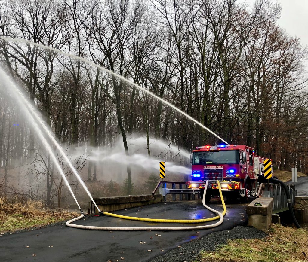 A fire truck sprays water from multiple hoses on a narrow road bridge surrounded by leafless trees. The truck has flashing lights, and the scene appears to be a training exercise or drill.