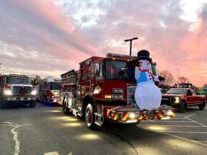 A red fire truck decorated with a large inflatable snowman and holiday lights is parked on a street at sunset, with other emergency vehicles visible in the background.