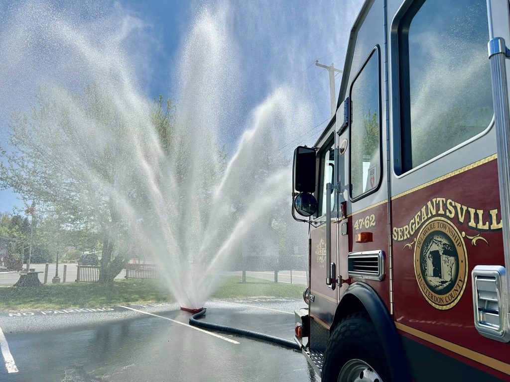 A fire truck labeled “Sergeantsville” sprays water in a large, fan-shaped arc across a sunny parking lot, creating a mist with trees and blue sky in the background.