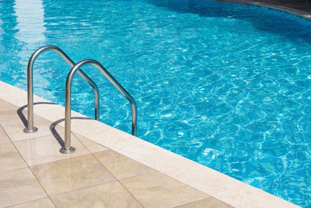 A stainless steel ladder leads into a clear blue swimming pool, with sunlight reflecting off the water. The pool is bordered by beige tiles.
